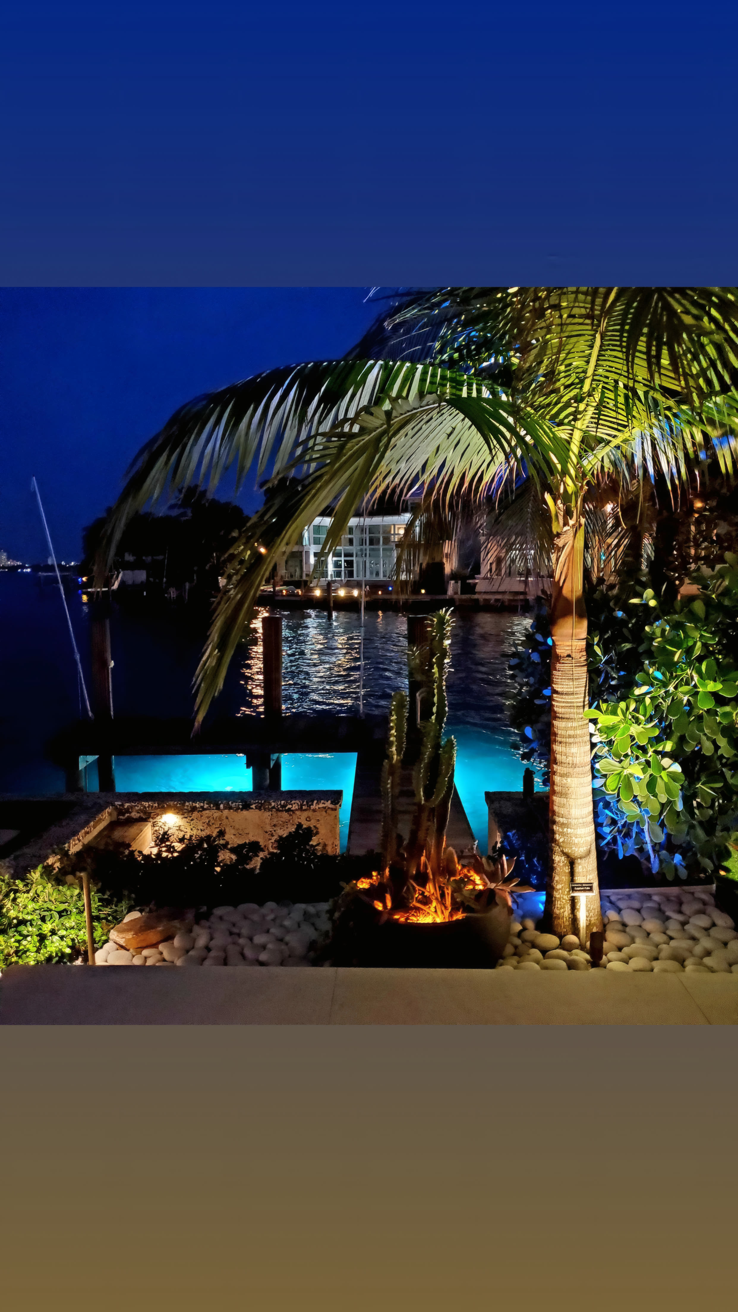 Waterfront tiki cabana at twilight with palm canopy, a small fire bowl, and uplit bay view